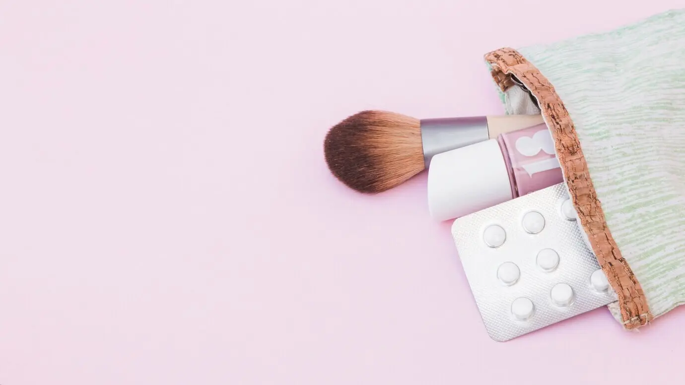 Vor pinkem Hintergrund ragen eine Nagellackflasche, ein Make-up-Pinsel und eine Blisterpackung mit Pillen aus einem Beutel.